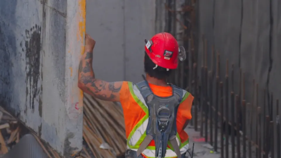 Coffreur au casque rouge, de dos, entre deux murs de béton pleine hauteur déjà coulés, tiges d'armature en attente le long de la paroi.
