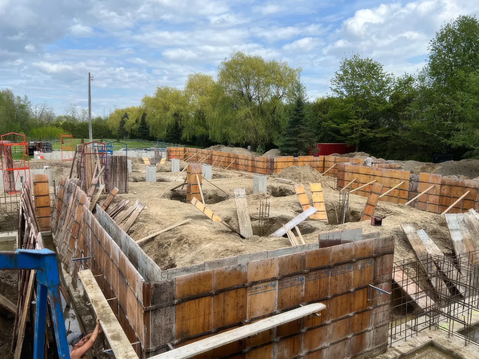 Coffrage de fondation déployé sur toute l'emprise d'un chantier agricole : panneaux de bois contreventés par des étais obliques, socles de poteaux et cages d'armature dans le sable, ciel bleu et rangée de saules à l'arrière.