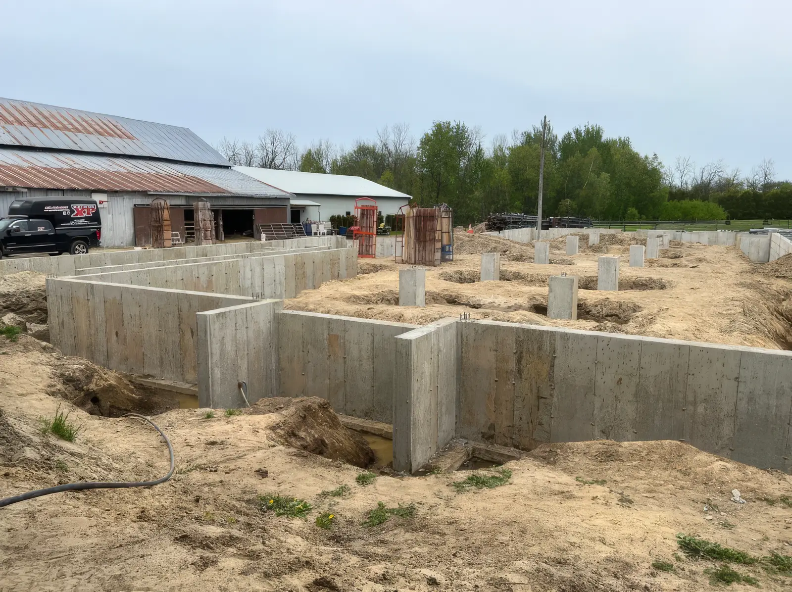 Fondation agricole coulée et décoffrée : longs murs de béton découpant plusieurs travées, socles de poteaux répartis dans le sable, grange de tôle et camion de l'entreprise à l'arrière.