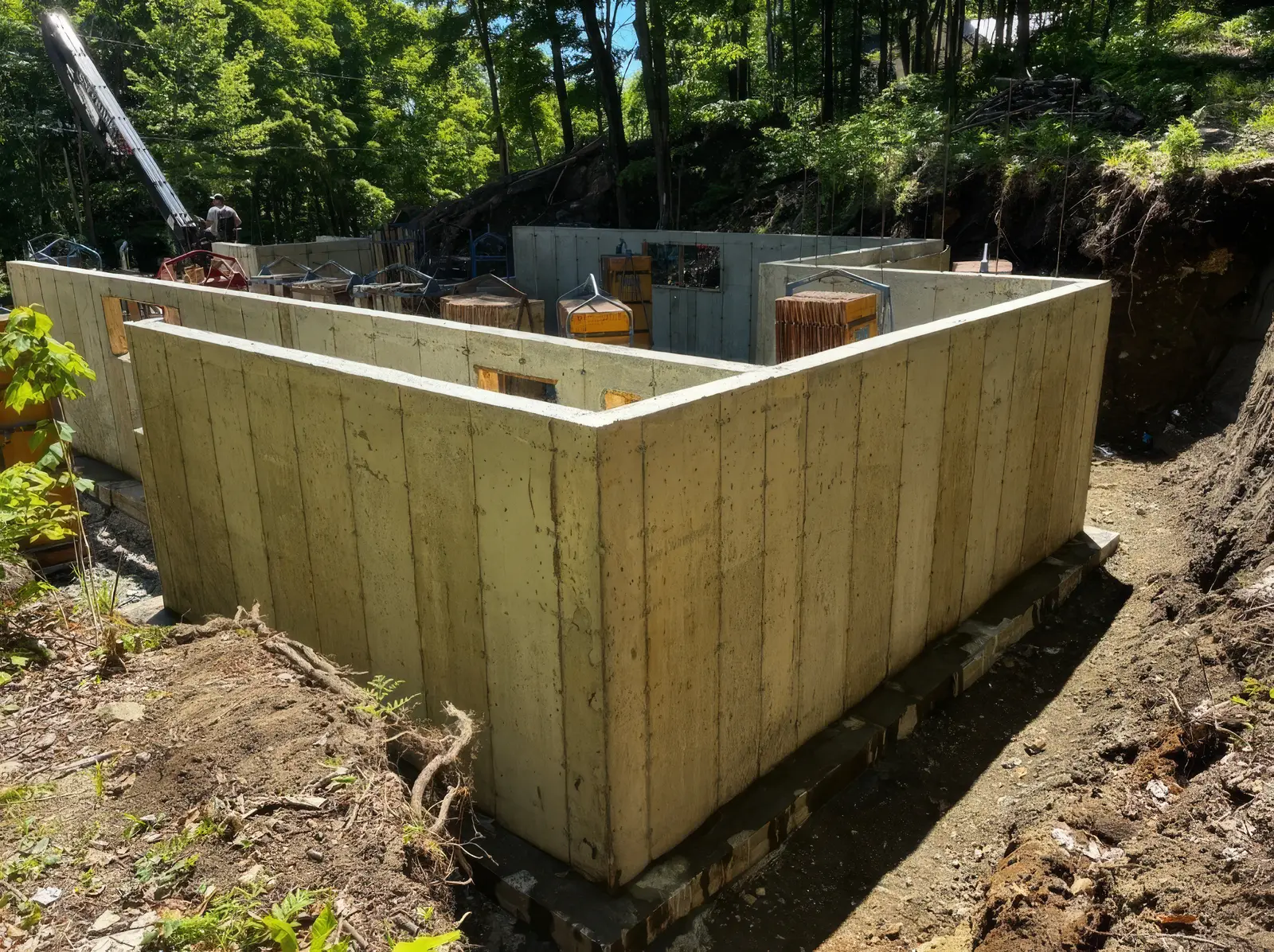 Fondation résidentielle entièrement décoffrée sur un terrain boisé en pente : murs de béton pleine hauteur formant un L, empreinte du bois de coffrage encore visible, panneaux et cages d'armature empilés à l'arrière, grue en bordure.