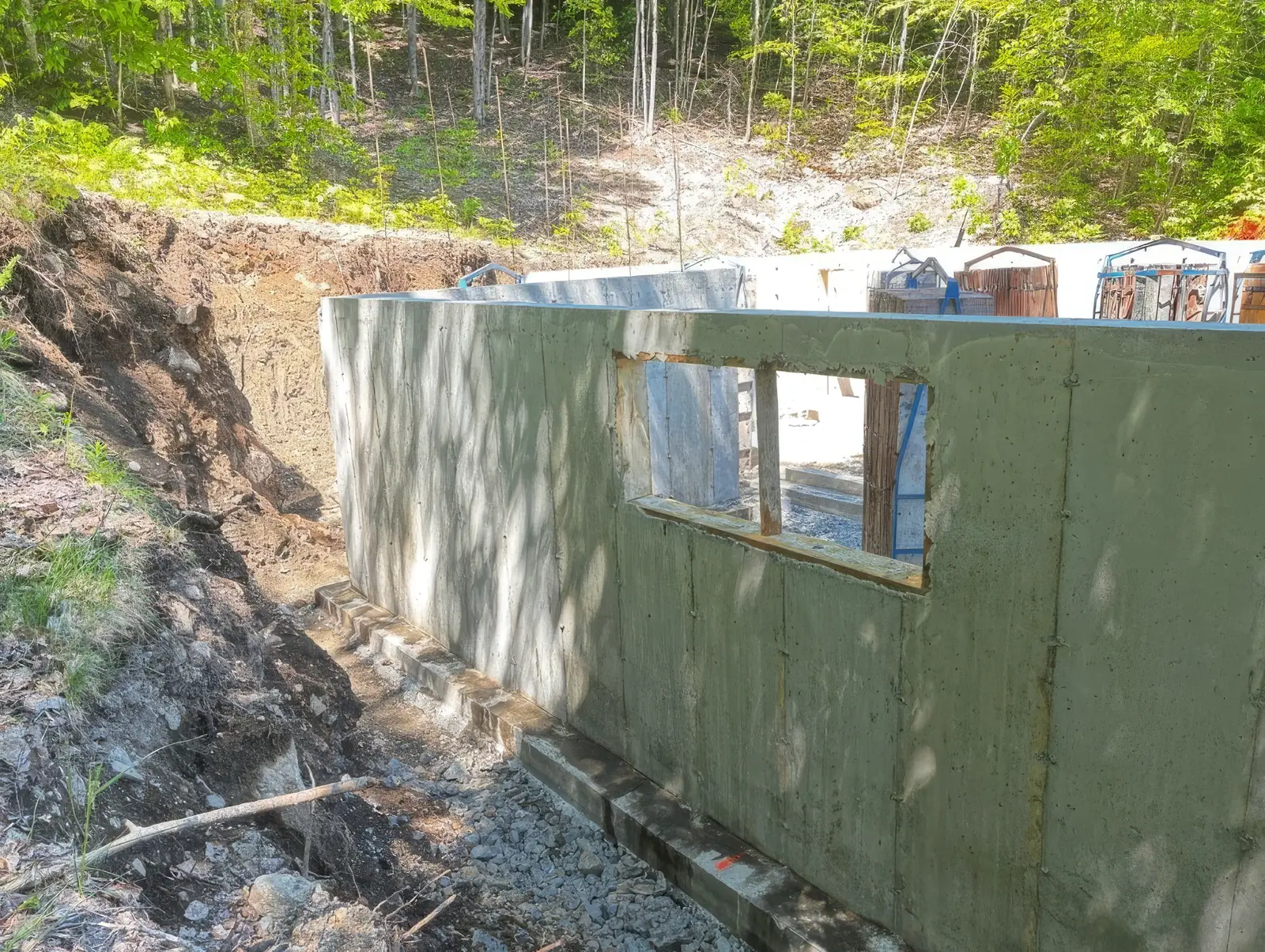 Mur de fondation décoffré longeant la paroi d'excavation en sous-bois, percé d'une ouverture de fenêtre encore encadrée de bois, semelle de béton visible au pied.