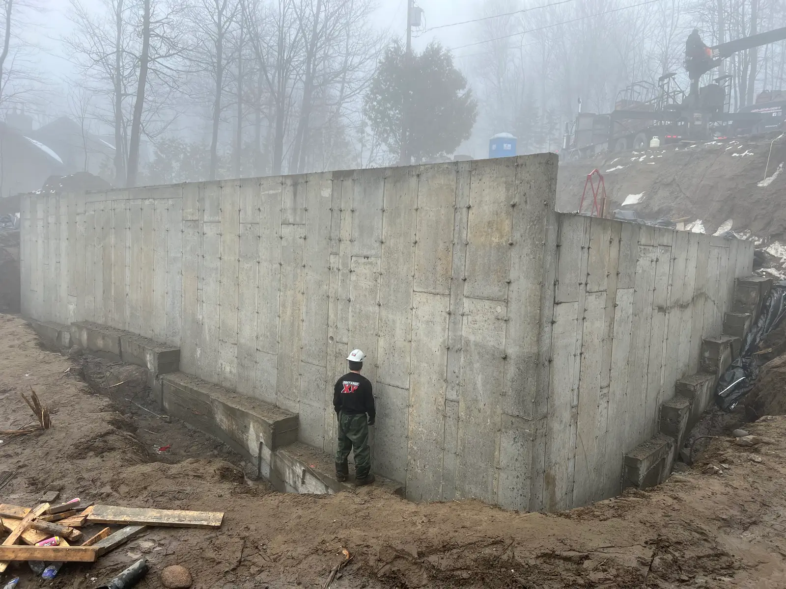 Mur de fondation décoffré de pleine hauteur dans la brume ; un travailleur debout à son pied donne l'échelle.