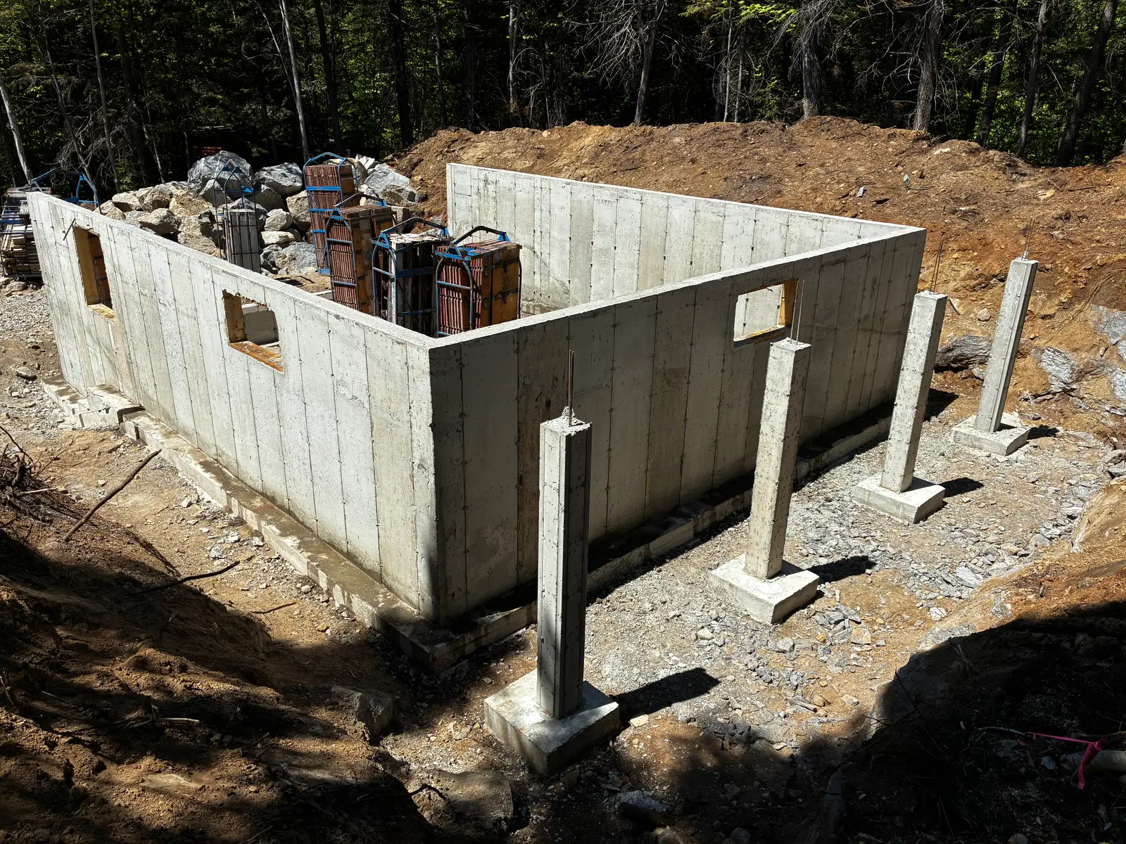 Fondation décoffrée en sous-bois : murs de béton pleine hauteur percés de deux ouvertures, poteaux d'acier posés sur leurs semelles, remblai de pierre.
