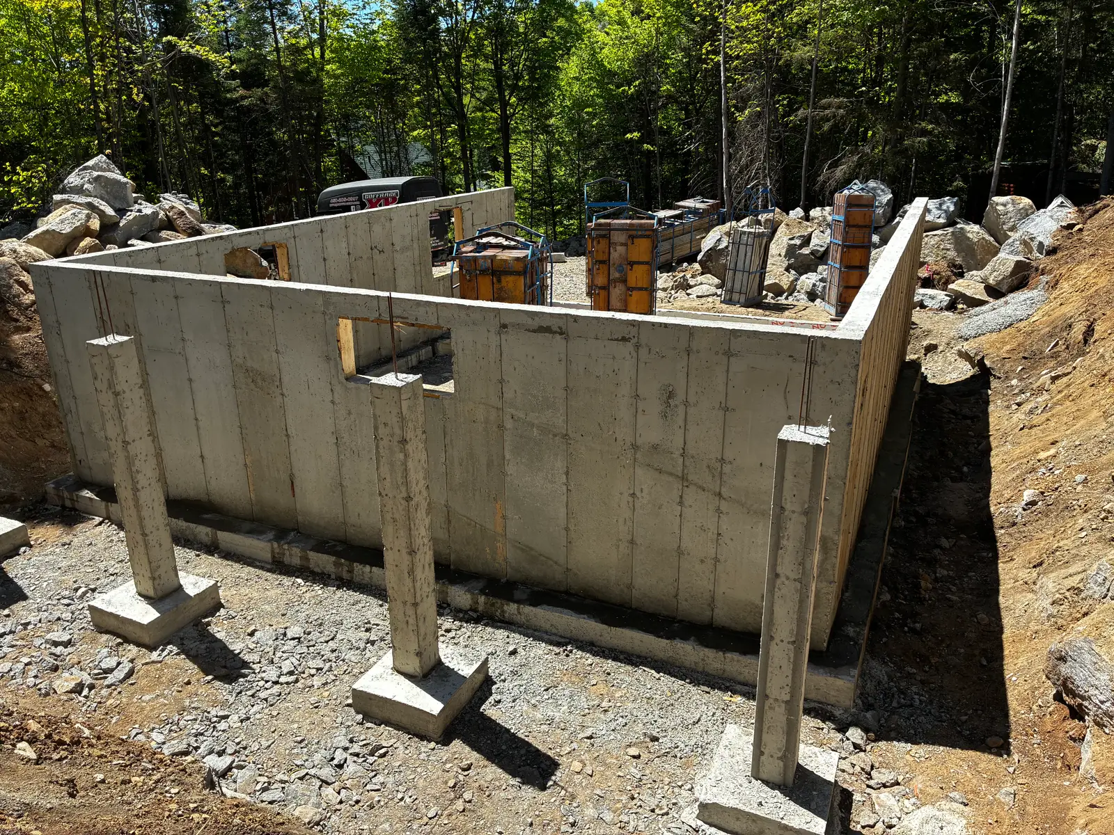 Sous-sol décoffré vu de l'intérieur : murs de béton, rangée de poteaux d'acier sur leurs semelles et amoncellement de roc derrière.