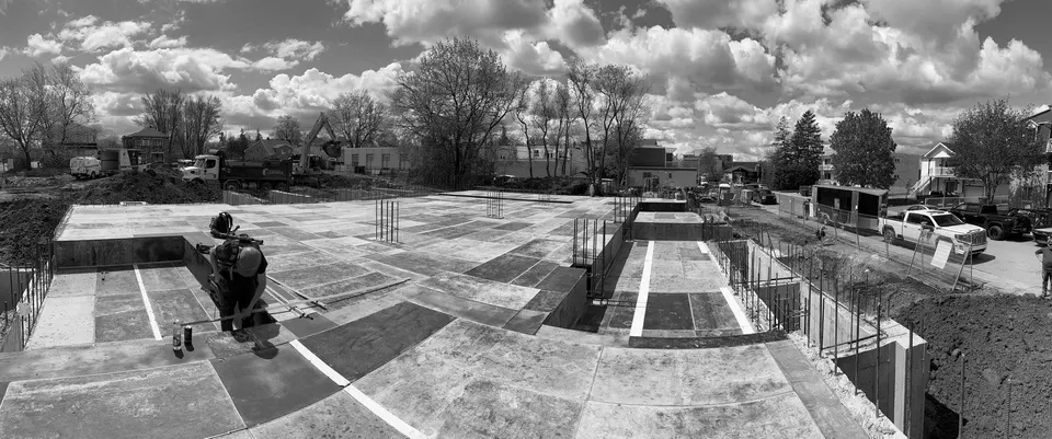 Panorama noir et blanc d'une dalle de béton terminée, coffrages de rive encore en place et quartier résidentiel derrière.
