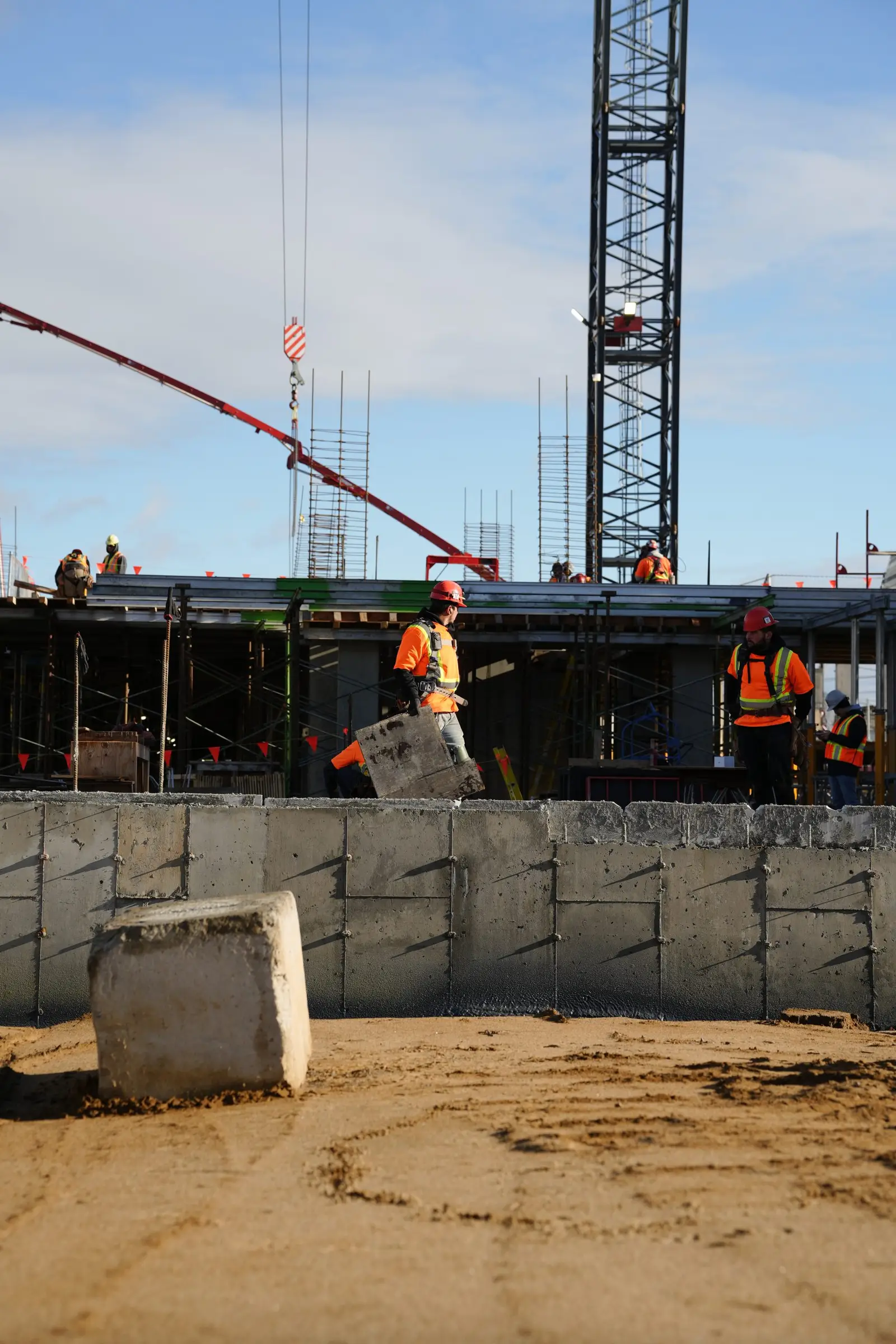 Mur de fondation décoffré au premier plan, marques d'attaches visibles dans le béton ; immeuble en construction et grue à tour à l'arrière.